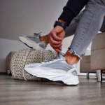 Person tying the laces of gray and white athletic sneakers, seated indoors with a chunky knit pouf nearby.