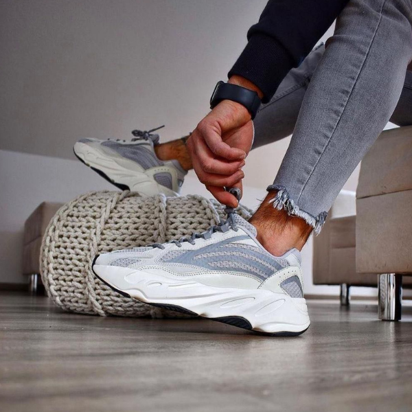 Person tying the laces of gray and white athletic sneakers, seated indoors with a chunky knit pouf nearby.