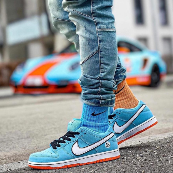 Close-up of blue Nike sneakers with white swoosh, orange sole, and blue socks, standing on a street with a colorful car in the background.