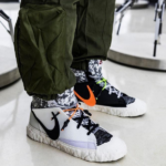Close-up of two Nike sneakers: the foreground white high-tops with black swoosh and orange laces, and a black pair with neon green laces in the background, worn with camo socks and olive cargo pants.
