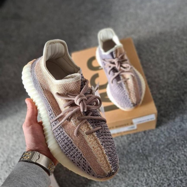 Pair of beige and lavender knit sneakers with thick cream soles, held in hand in front of their box.