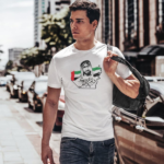 Young man walking on a city street, carrying a black backpack over his shoulder and wearing a white T‑shirt with a graphic featuring a portrait and a flag (green, white, red).