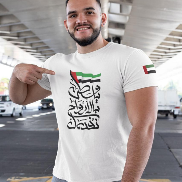 Untitleddesign_85_74cfbbae-9302-43f0-82a9-31df7d6400e7 | UAE SPORT STORE Man in a white t-shirt pointing to bold Arabic calligraphy text on the chest, with UAE flag colors above it and a flag on the sleeve, in an outdoor setting.