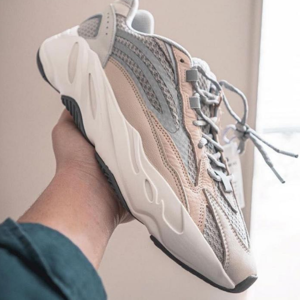 Person holding a beige and gray chunky sneaker with a thick white sole near the camera on a light background, shoe details visible