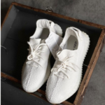 Pair of white knit sneakers with laces, placed in an open wooden crate; Yeezy branding visible inside.