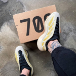 Person wearing white, yellow, and black sneakers stands on a concrete floor with a cardboard sign reading 700 partially underfoot.
