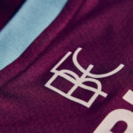 Close-up of maroon sports jersey fabric with a light blue stripe and a white partial logo/letter visible.