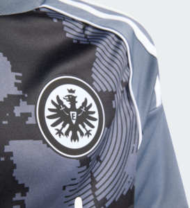 Close-up of a dark blue athletic jersey with a circular black-and-white crest featuring a crowned eagle; digital camouflage pattern in the background and white piping along the shoulder.