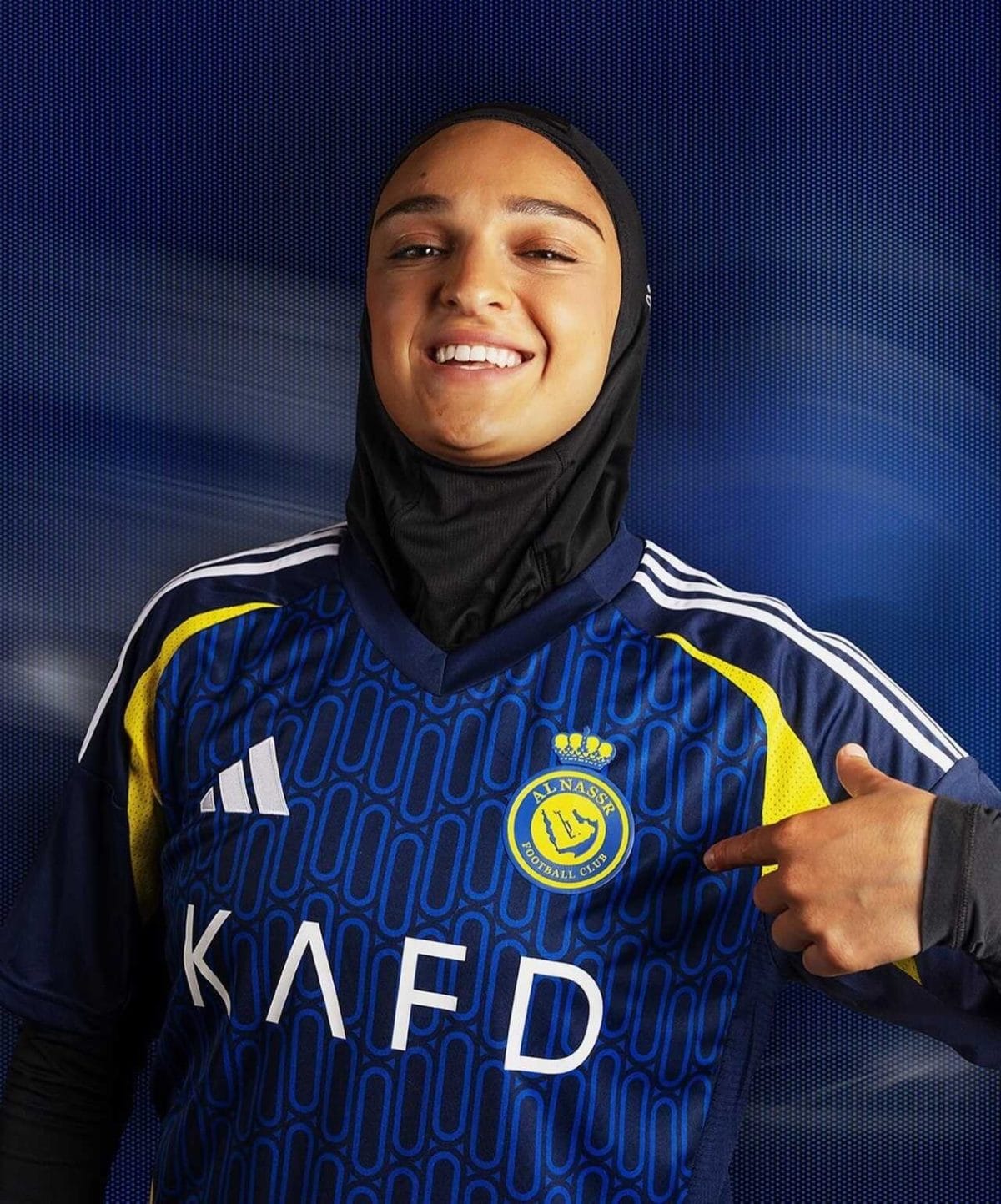 Smiling woman in a blue football jersey with a hijab, pointing to the team crest on her chest.