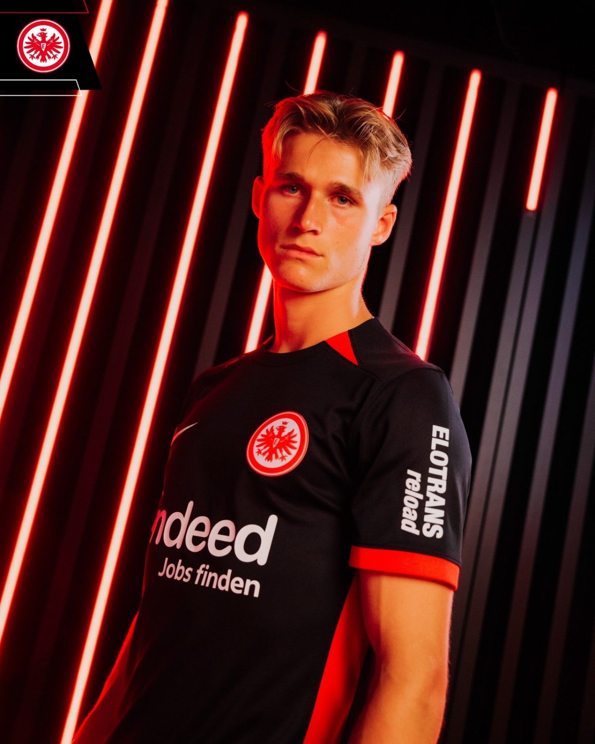 Young man wearing a black Eintracht Frankfurt jersey stands against red neon light stripes, with team crest visible on the chest and the word 'indeed' on the front.