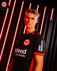 Va4dbVmZXAQJVGn | UAE SPORT STORE Young man wearing a black Eintracht Frankfurt jersey stands against red neon light stripes, with team crest visible on the chest and the word 'indeed' on the front.