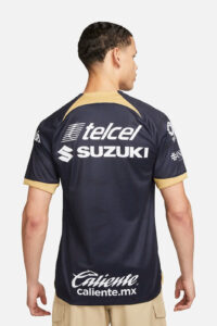 Back view of a man wearing a navy jersey with tan trim, showing Telcel, Suzuki, and Caliente.mx logos on the back.