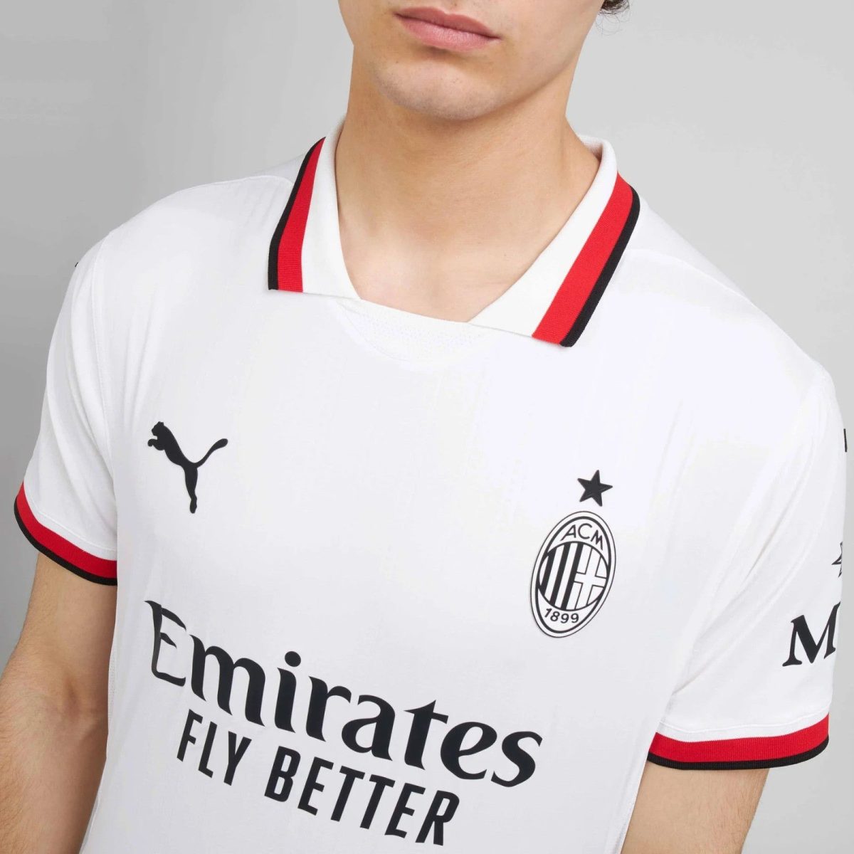 Partial view of a person wearing a white AC Milan football jersey with a red and black striped collar, featuring the Puma logo, club crest, and 'Emirates Fly Better' text.