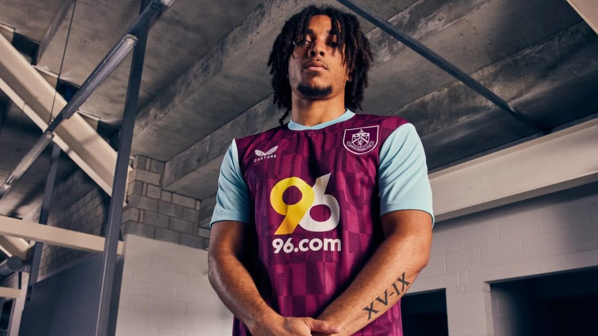Athlete in a maroon and light-blue soccer jersey with 96.com sponsor, standing in an industrial indoor setting, arms folded reveal Roman numeral tattoo XV-IX on forearm.