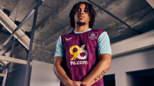 Athlete in a maroon and light-blue soccer jersey with 96.com sponsor, standing in an industrial indoor setting, arms folded reveal Roman numeral tattoo XV-IX on forearm.