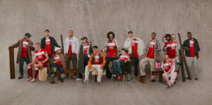 Diverse group of people in red-and-white UNIBET shirts posing together in a studio setting, some seated or leaning on props like benches and planks to create a casual team photo.
