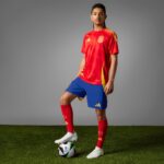 Soccer player in red jersey and blue shorts stands on grass with a ball at his foot against a gray studio backdrop