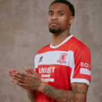 Man in a red-and-white football jersey with a crest, looking at the camera with hands clasped in front, tattoos visible on arms and neck.