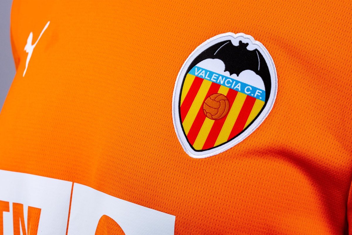 Valencia CF crest on an orange jersey featuring red-yellow stripes, a blue name banner, and a black bat silhouette in the top.