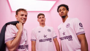 Three young men in pink room wearing white soccer jerseys with Hollywood Bets logo, posing for a team photo.