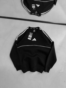 Two black Adidas sweatshirts with white stripes laid on a cracked concrete floor, tags still attached to the tops.