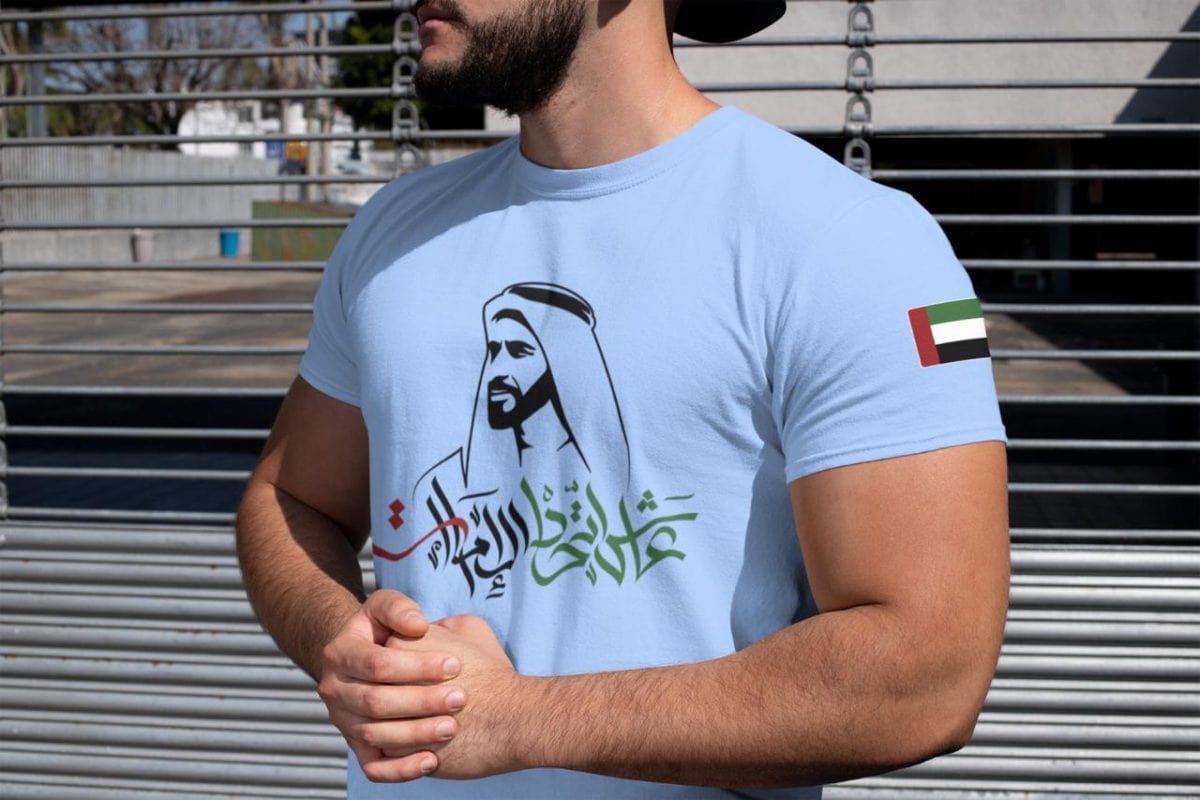 Man wearing a light blue T-shirt with Arabic calligraphy and a headscarf silhouette; UAE flag patch on the sleeve, standing by a metal fence.