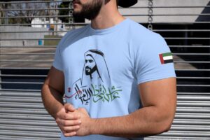 Man wearing a light blue T-shirt with Arabic calligraphy and a headscarf silhouette; UAE flag patch on the sleeve, standing by a metal fence.