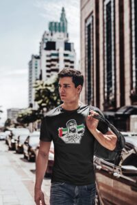Man walking down a city street wearing a black T-shirt with a Arabic-script graphic and carrying a backpack over his shoulder.