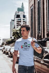 Young man walking down a city street wearing a light blue graphic T-shirt and a black backpack over his shoulder.