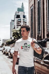 Young man with short dark hair walking on a sunny urban street, carrying a backpack over his shoulder, in a white graphic tee.