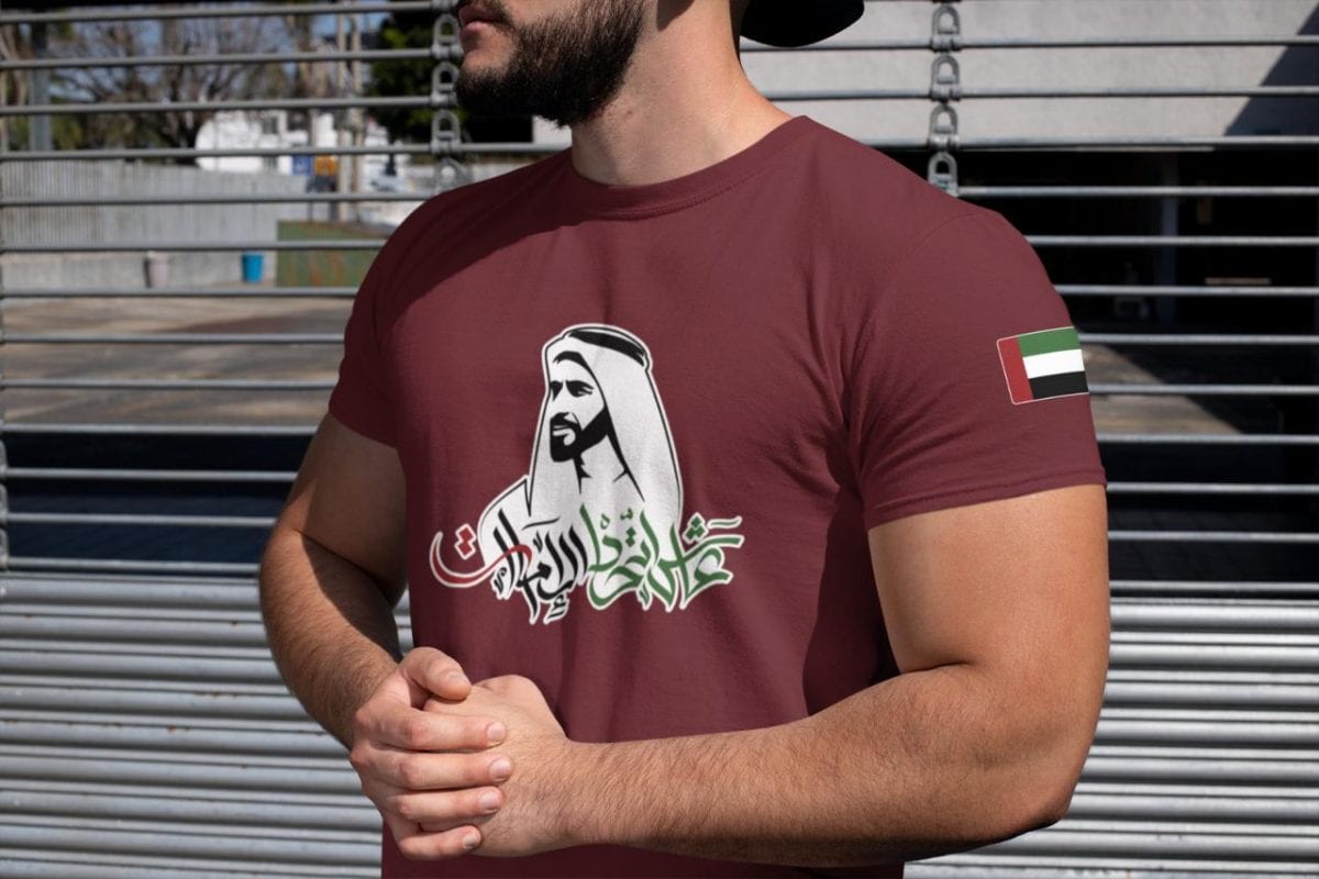 Man wearing a maroon T-shirt with Arabic calligraphy and a white-robe illustration on the chest; UAE flag on the sleeve.