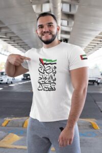 WhatsAppImage2020-11-24at7.53.55PM_34159f60-c818-422f-ba30-62fbc0f820b9 | UAE SPORT STORE Smiling man in a white T-shirt with Arabic script and a UAE flag patch on the sleeve, pointing to the logo on his chest under a highway overpass.