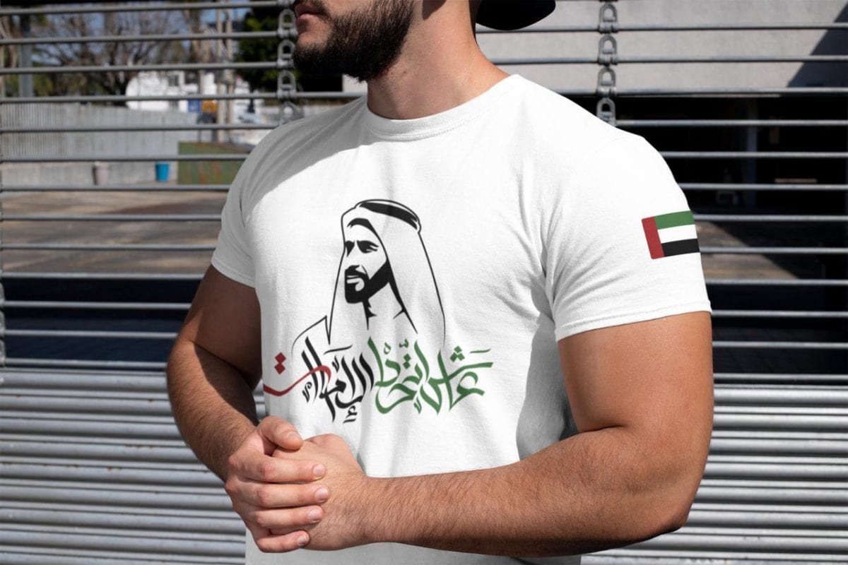 Man wearing a white T-shirt with green Arabic script and a black silhouette of a man in traditional dress; UAE flag patch on the sleeve behind a metal fence backdrop.