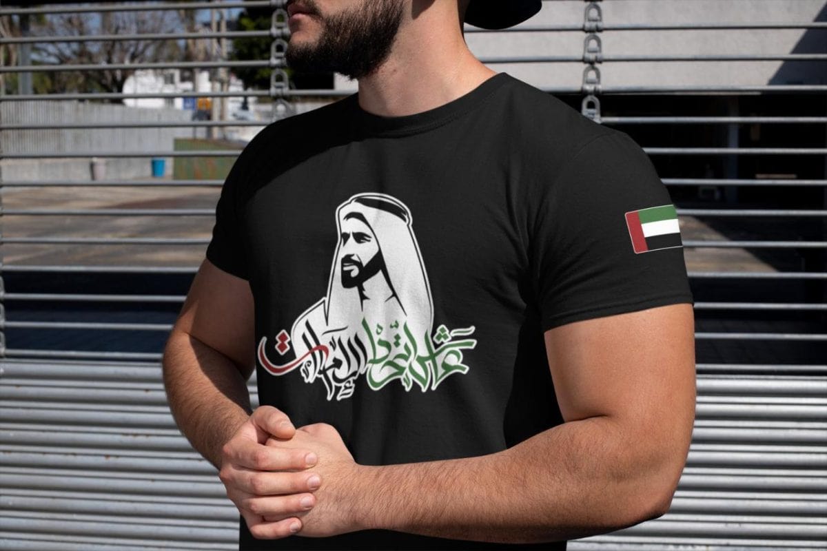 Man wearing a black T-shirt with Arabic calligraphy design and a UAE flag on the sleeve, outdoors by a metal fence.