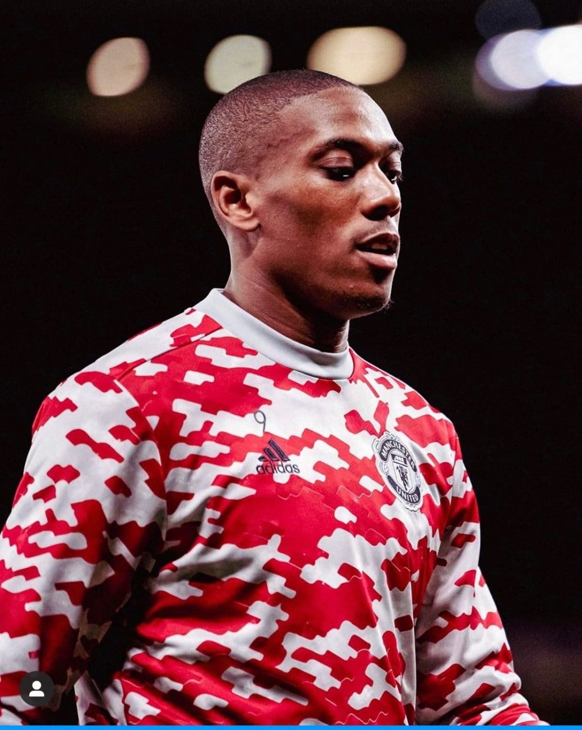 Portrait of a man in a red and white camouflage Manchester United jersey, looking to the right, with a dark blurred background and stadium lights in the distance.