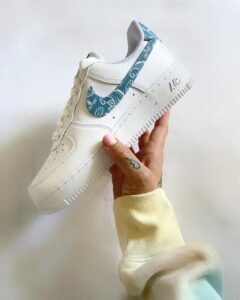 WhatsAppImage2022-09-18at09.15.49_90e4fcdf-59b2-4a51-ab9c-9e75ef5a7c3a | UAE SPORT STORE White Nike Air Force 1 sneaker with a light blue patterned swoosh held in a hand against a pale background.