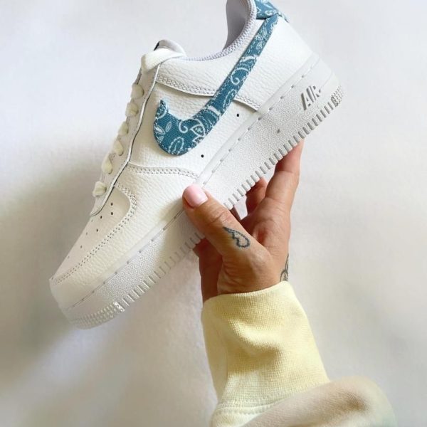 White Nike Air Force 1 sneaker with a light blue patterned swoosh held in a hand against a pale background.