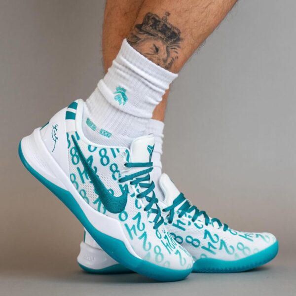 White and teal basketball sneakers with a number-print pattern, teal swoosh, and white socks on a tattooed lower leg; dynamic low-angle shot.