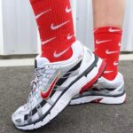 Runner wearing white, gray, and black Nike running shoes with a red Nike swoosh and red socks standing on pavement.