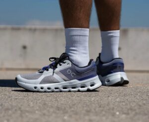 Close-up of gray and blue running shoes on a person wearing white socks, standing on paved ground by a concrete barrier.