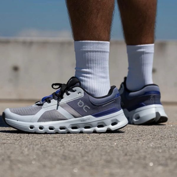 Close-up of gray and blue running shoes on a person wearing white socks, standing on paved ground by a concrete barrier.