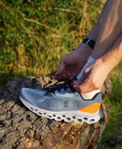 Person tying the laces of a light blue running shoe with an orange heel on a rocky surface outdoors