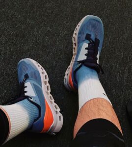 Person wearing blue athletic sneakers with white soles and orange accents, standing on a dark textured surface in white socks.