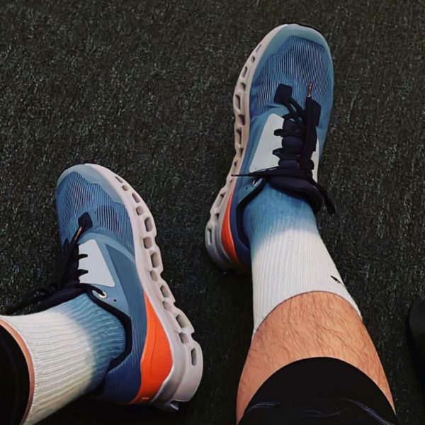 Person wearing blue athletic sneakers with white soles and orange accents, standing on a dark textured surface in white socks.