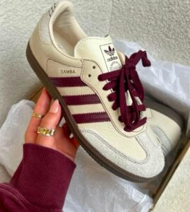 Cream Adidas Samba sneaker with burgundy stripes and laces being held in a hand over a box.