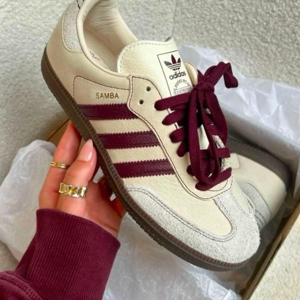 Cream Adidas Samba sneaker with burgundy stripes and laces being held in a hand over a box.