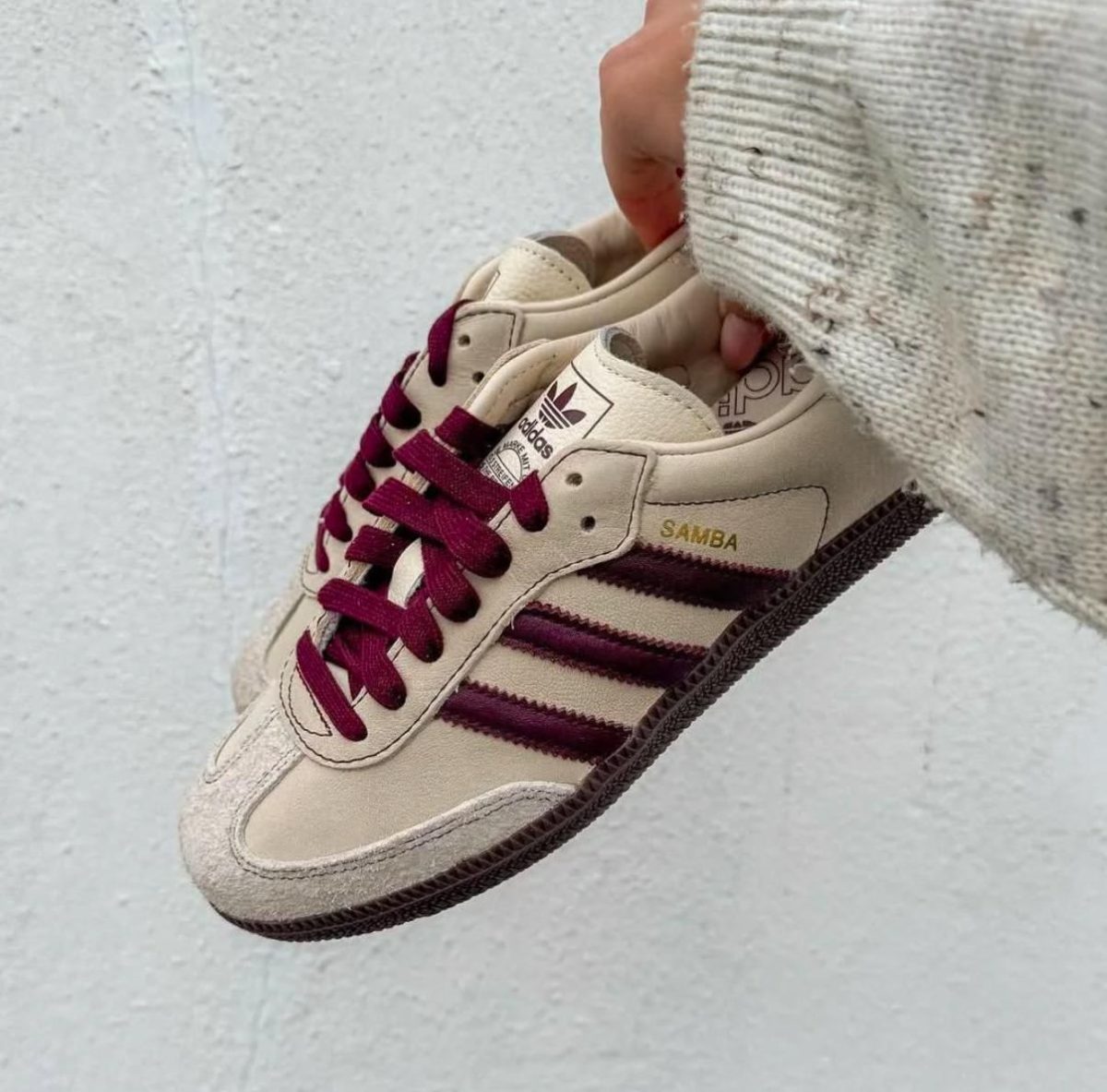 Beige Adidas Samba sneaker with burgundy laces held by a hand against a light concrete wall