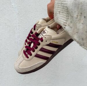 Beige Adidas Samba sneaker with burgundy laces held by a hand against a light concrete wall
