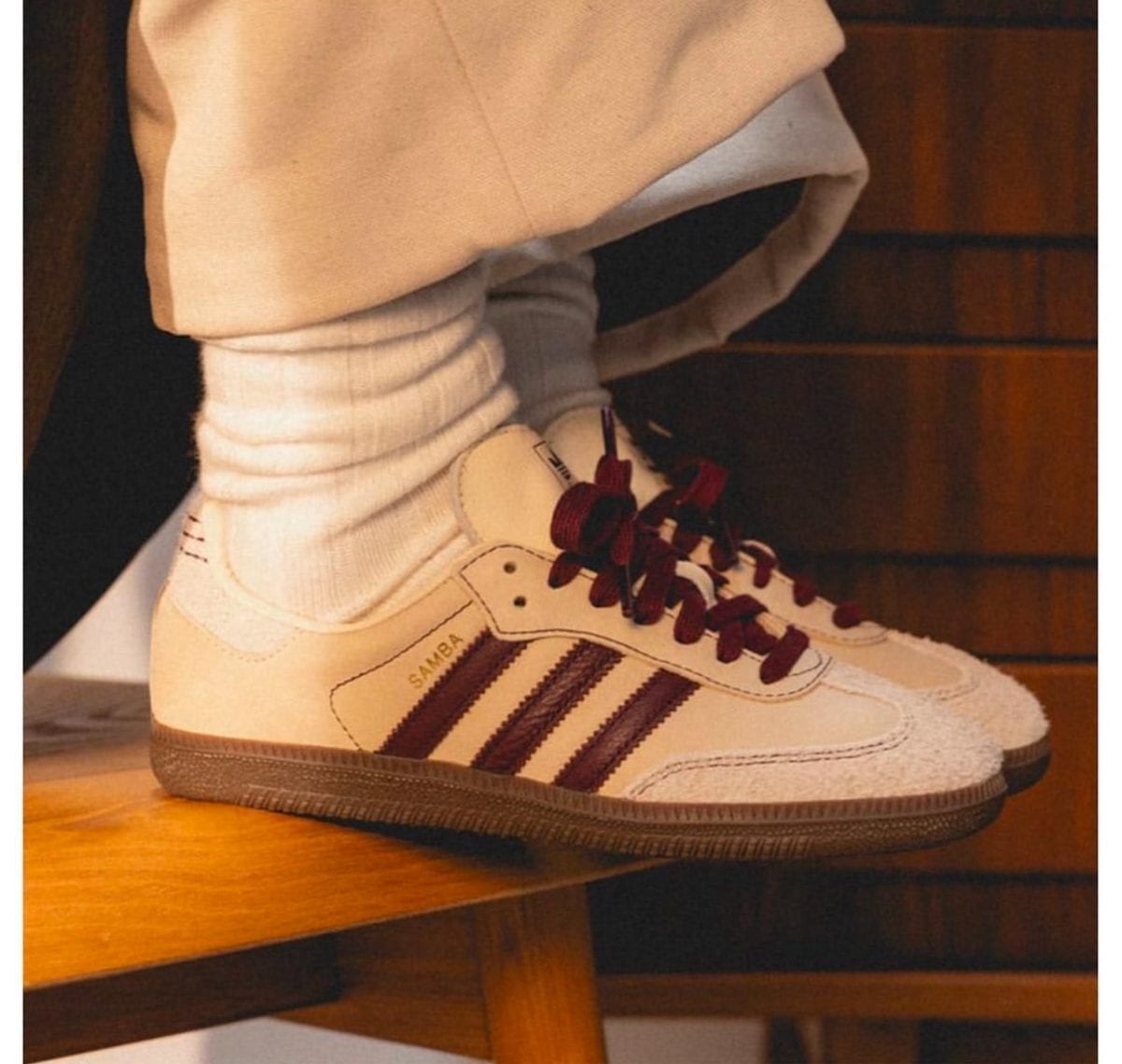 White Adidas Samba sneakers with burgundy laces, worn with white socks on a wooden ledge.