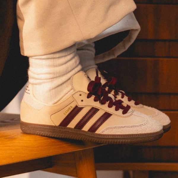 White Adidas Samba sneakers with burgundy laces, worn with white socks on a wooden ledge.